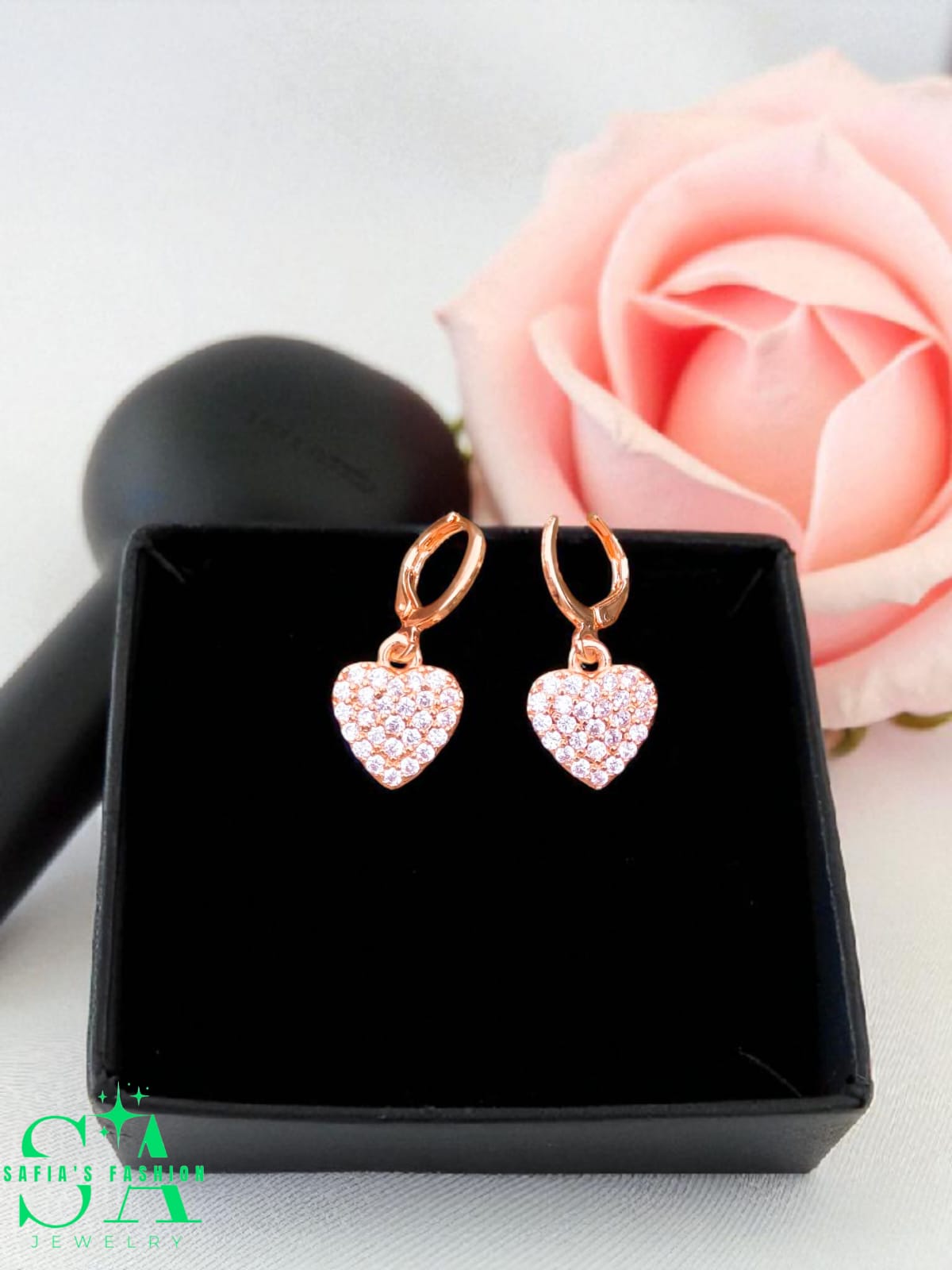 Heart-shaped earrings with rose gold finish and clear stones in a black box with a pink rose in the background.